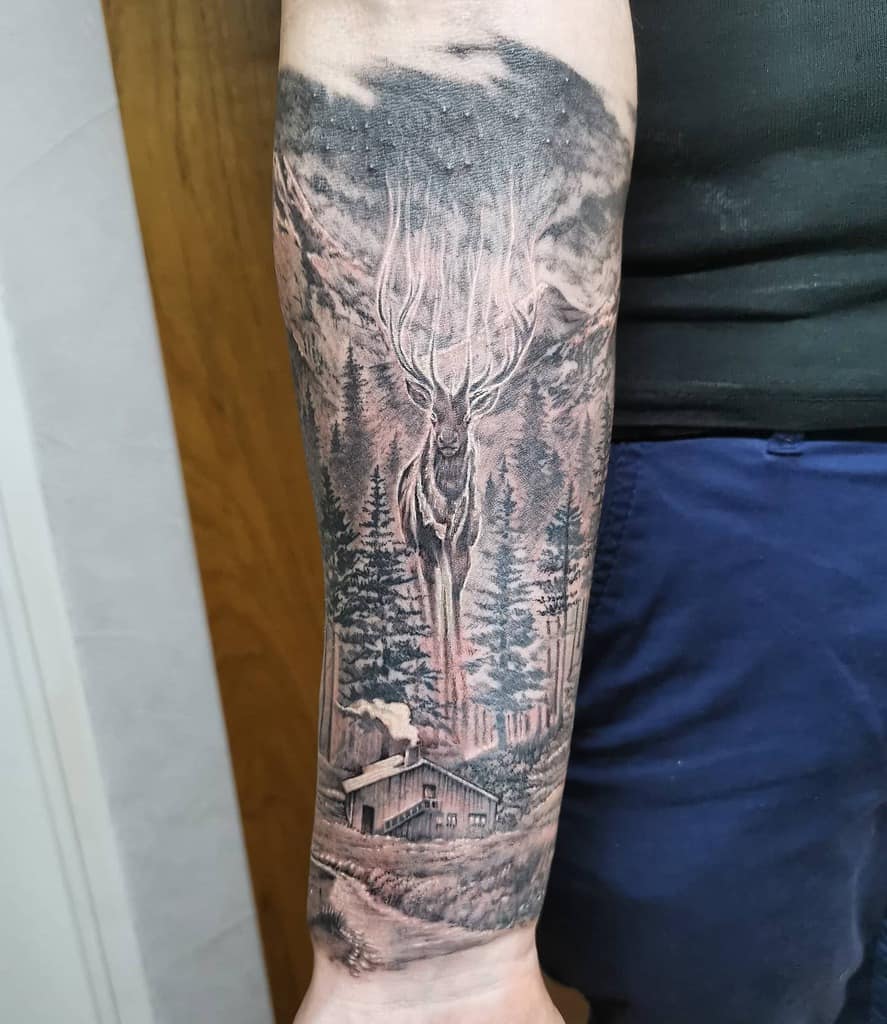 Forearm tattoo of a deer silhouette in a forest with tree outlines and a mountain cabin scene