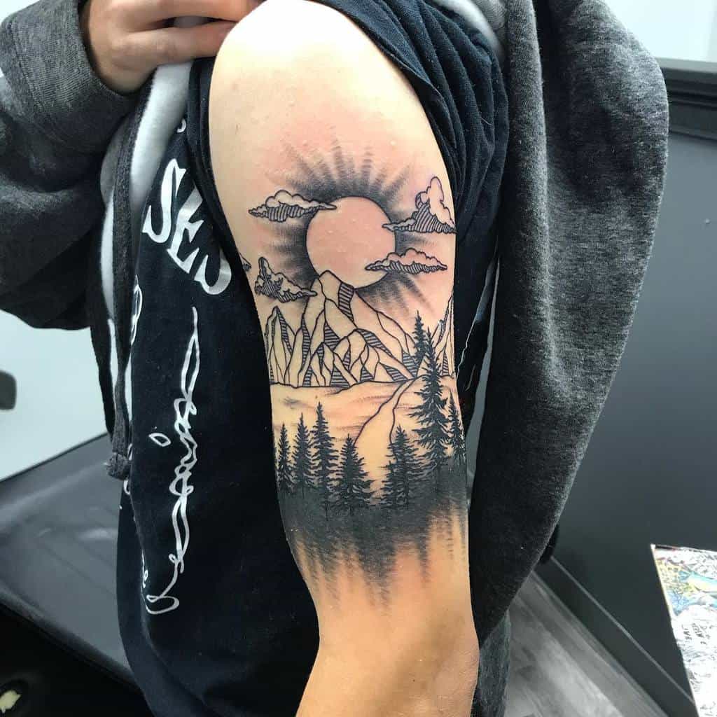 Upper arm tattoo featuring a striking silhouette of mountains, trees, and the sun with clouds