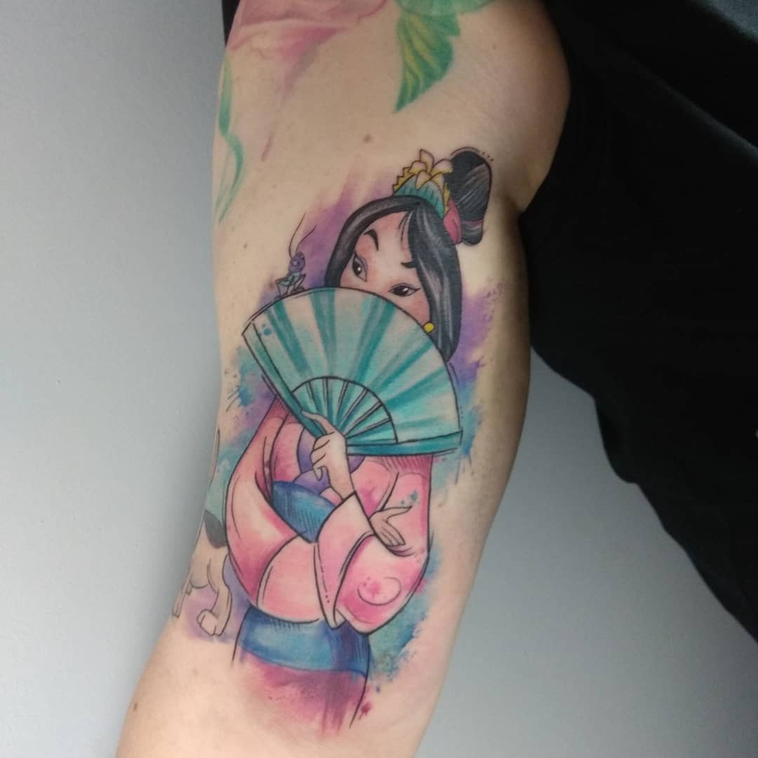 Tattoo of Mulan in traditional attire holding a fan on an upper arm