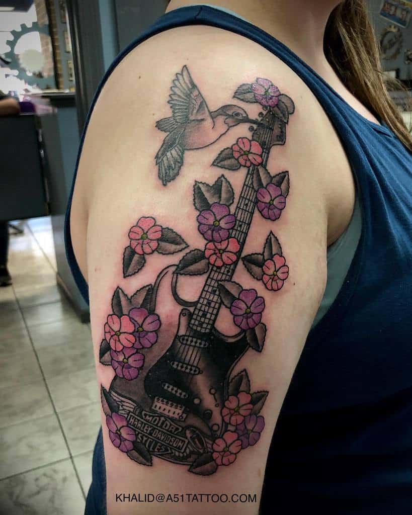 A tattoo of a guitar entwined with flowers and leaves, with a bird nearby, on an upper arm