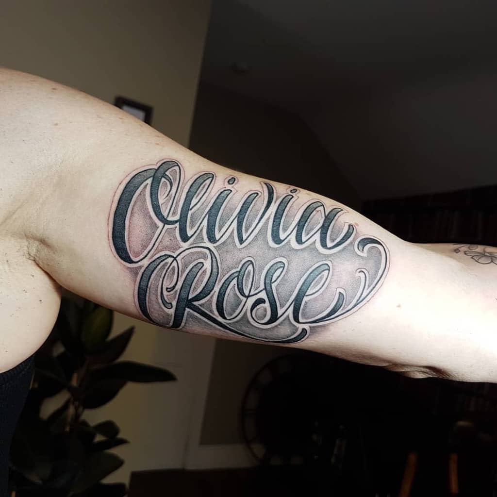 Bold inner arm tattoo with the name 'Olivia Rose' in elegant cursive script, shaded with black and grey for depth