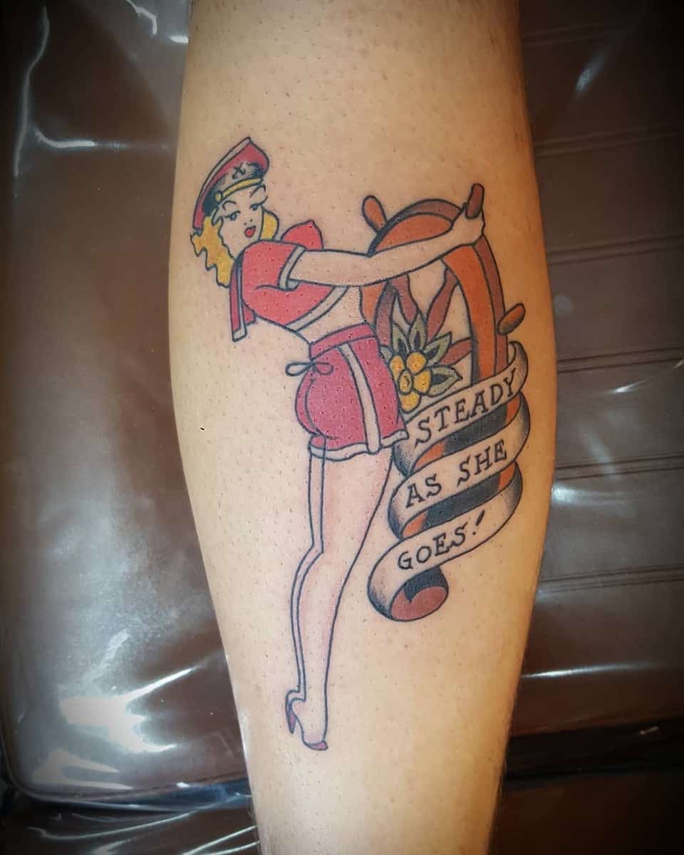 Tattoo of a pin-up sailor girl holding a ship's wheel with text "STEADY AS SHE GOES!" on a calf