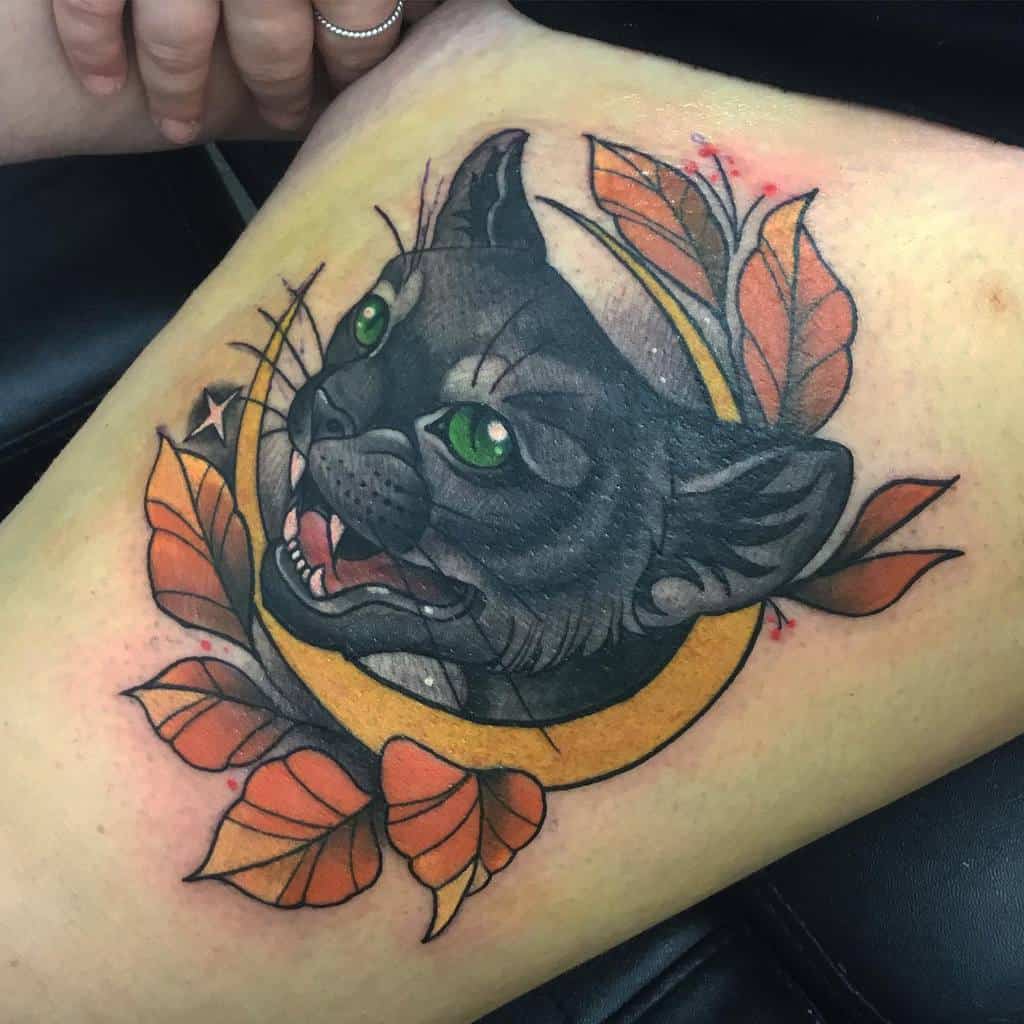 Tattoo of a sleek black cat with piercing green eyes, surrounded by vibrant autumn leaves and an enchanting orange crescent