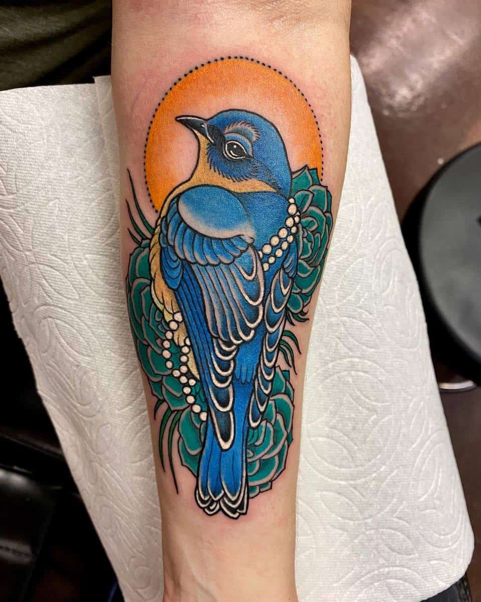 A colorful tattoo of a blue bird with a halo and surrounding green leaves on an arm