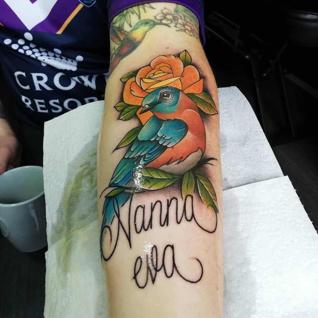 A colorful bird tattoo with an orange rose and the text "Nanna Eva" on a forearm