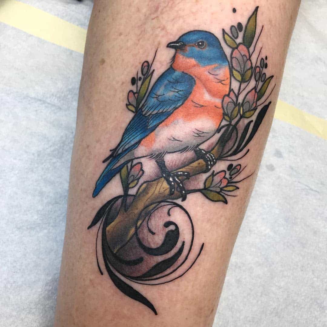 Tattoo of a blue and orange bird perched on a branch with flowers and leaves on an individual's arm