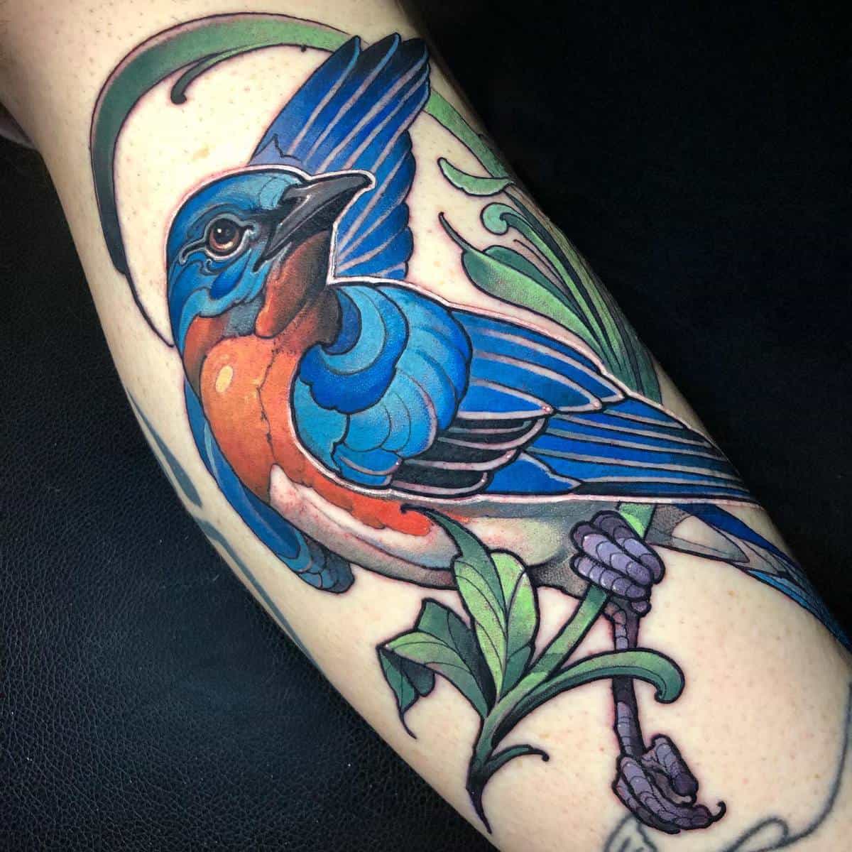 A vibrant tattoo of a blue and orange bird perched on a green branch on an arm