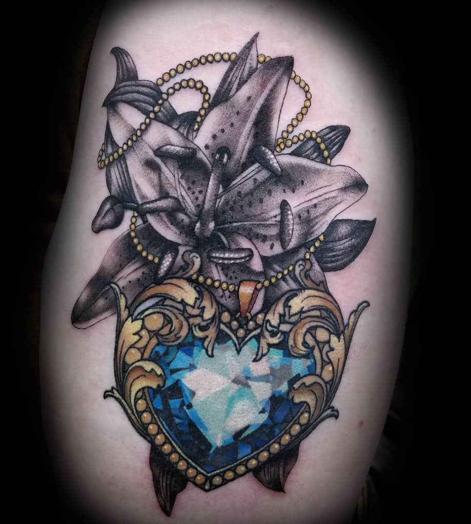 Ornate tattoo featuring black and grey lilies with golden beads, paired with a detailed blue heart-shaped gem in a baroque-style frame