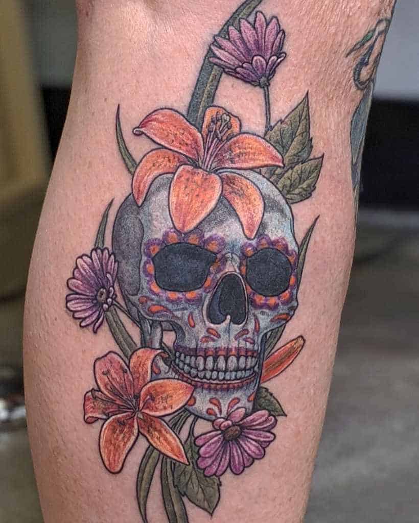 Colorful tattoo of a sugar skull adorned with orange lilies and purple flowers, blending Día de los Muertos symbolism with floral beauty