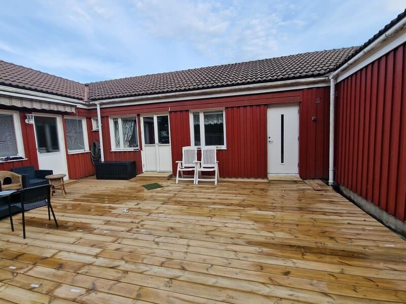 A wooden patio with white chairs, set against a red house with white trim and a tiled roof under a blue sky