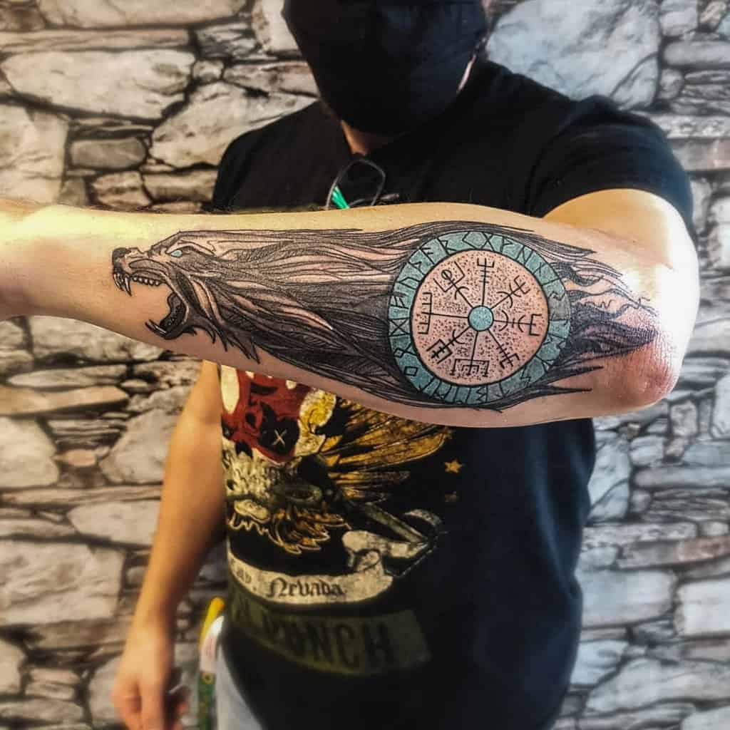 A Nordic forearm tattoo featuring a fierce wolf's head and a prominent Vegvisir symbol with blue accents and runic inscriptions