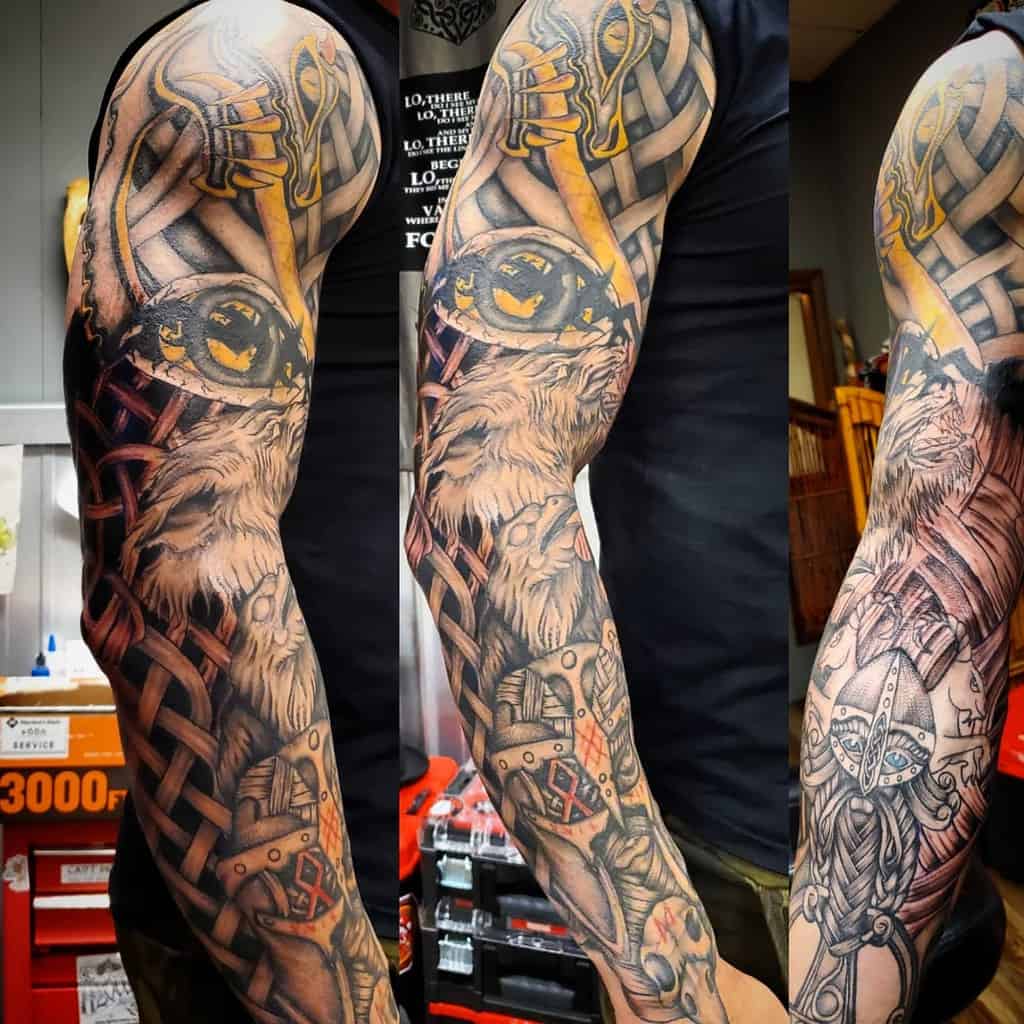 A full-sleeve tattoo featuring a detailed Viking warrior, an axe, a wolf, knotwork, and armor elements with bold accents of yellow and red