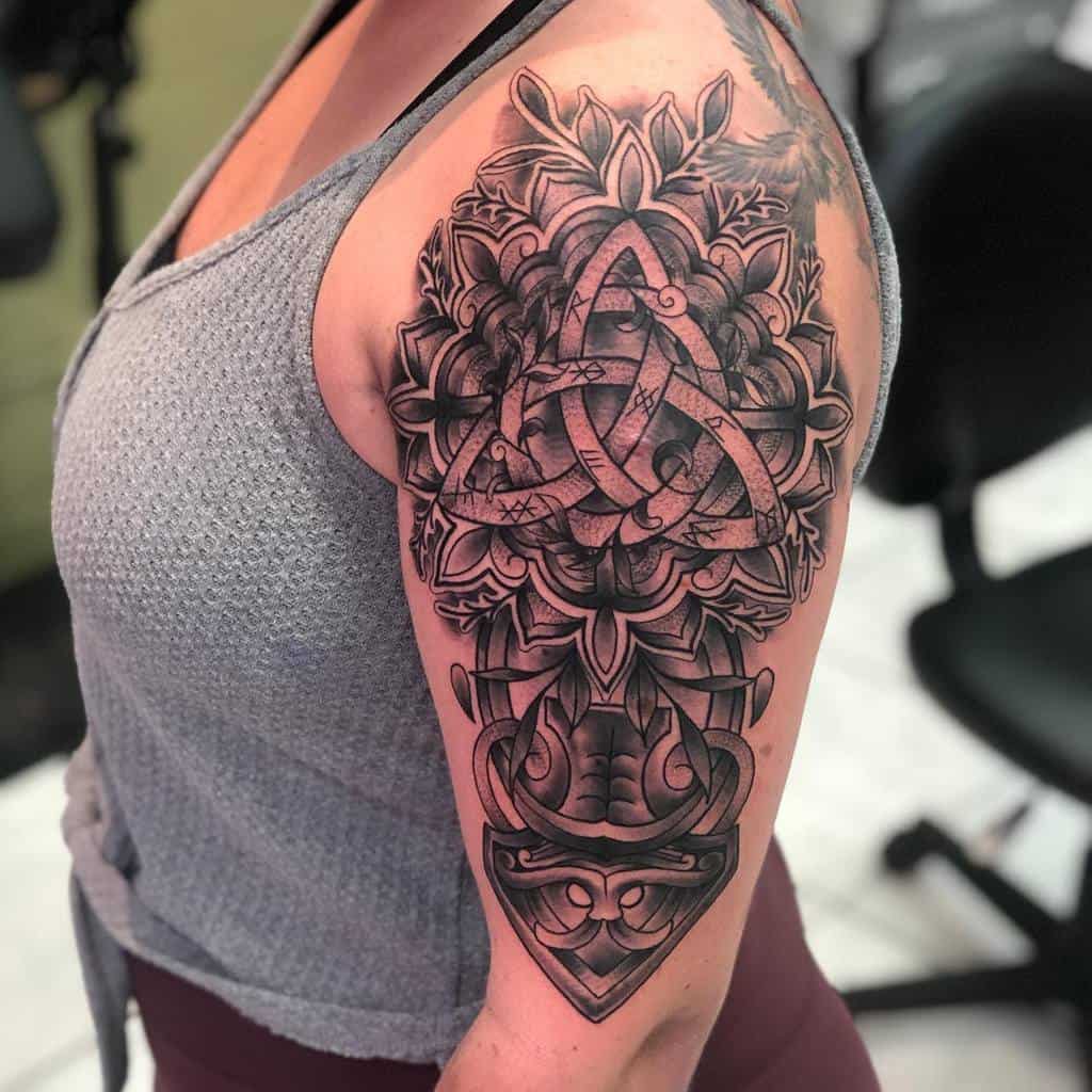 A Nordic shoulder tattoo featuring a detailed Valknut symbol, surrounded by intricate knotwork and floral patterns, with bold shading and a Viking helmet at the bottom
