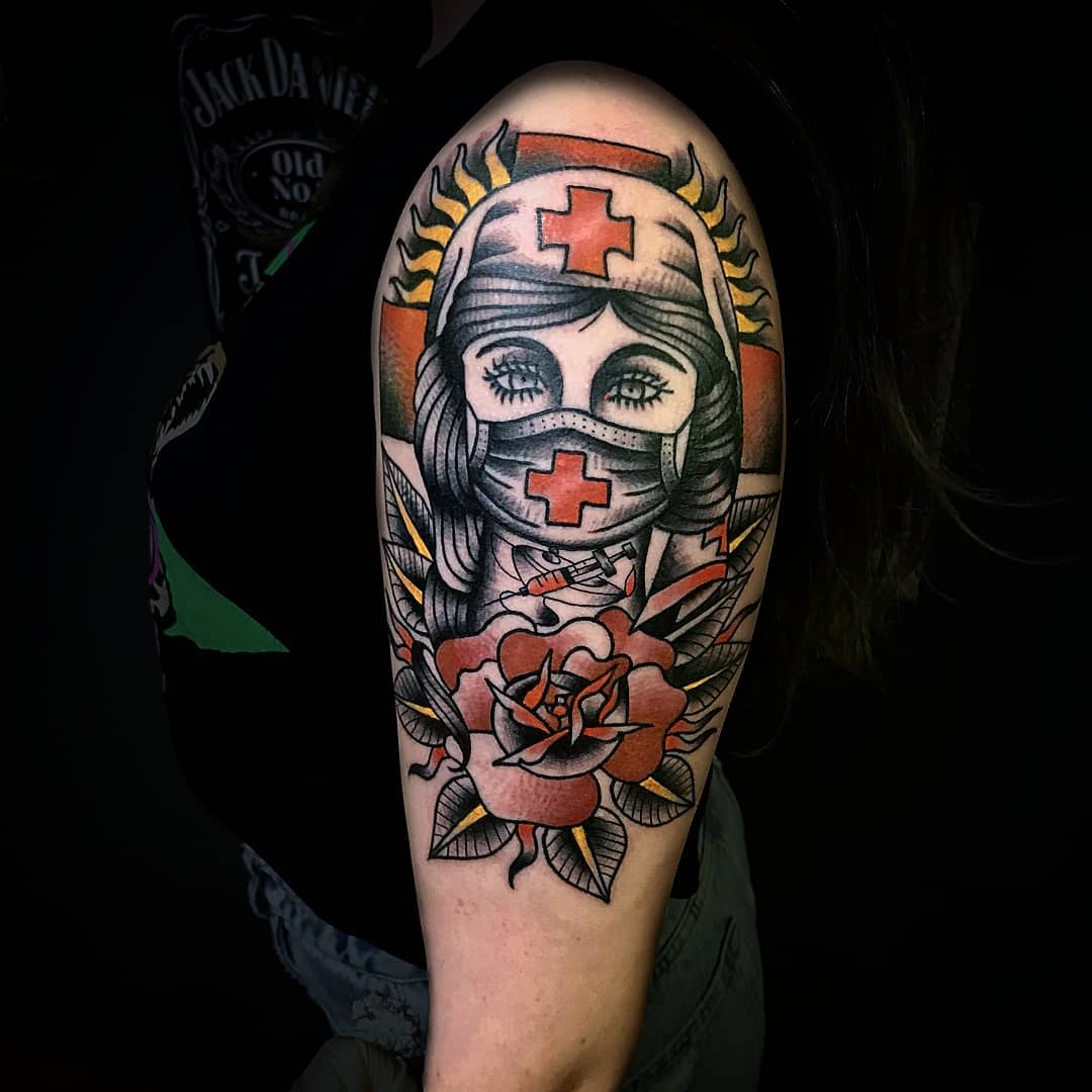 Tattoo of a nurse in a mask with a red cross, surrounded by flowers on an upper arm