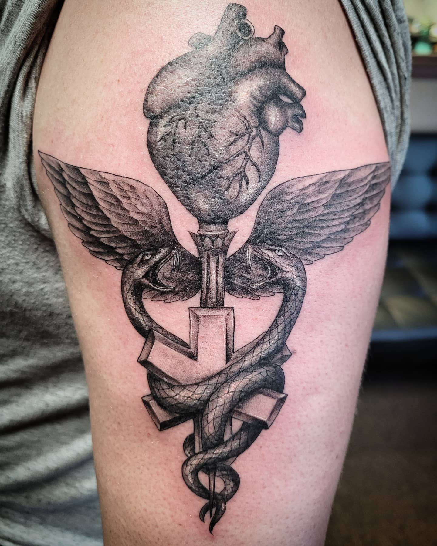 Tattoo of a heart above a staff with two snakes and wings on an upper arm