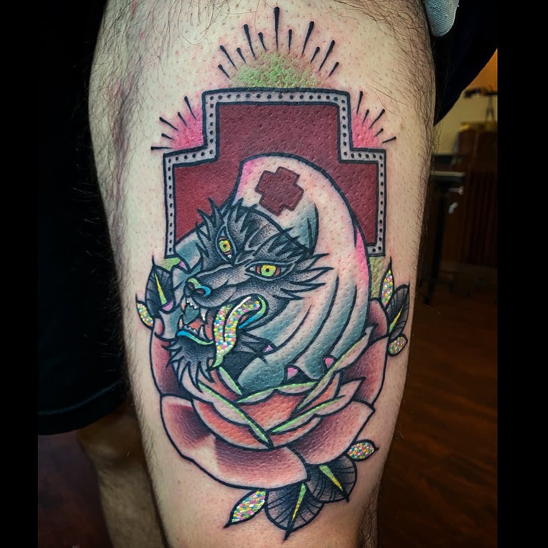 Colorful tattoo of a wolf head inside a nurse cap, atop a rose, on a leg