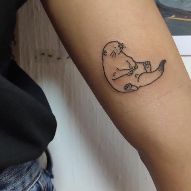 A minimalistic tattoo of an otter sits on an arm, paws up for a charming touch