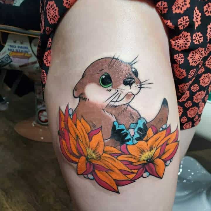 Colorful tattoo of a cartoon otter holding blue gems, with orange flowers, on a thigh