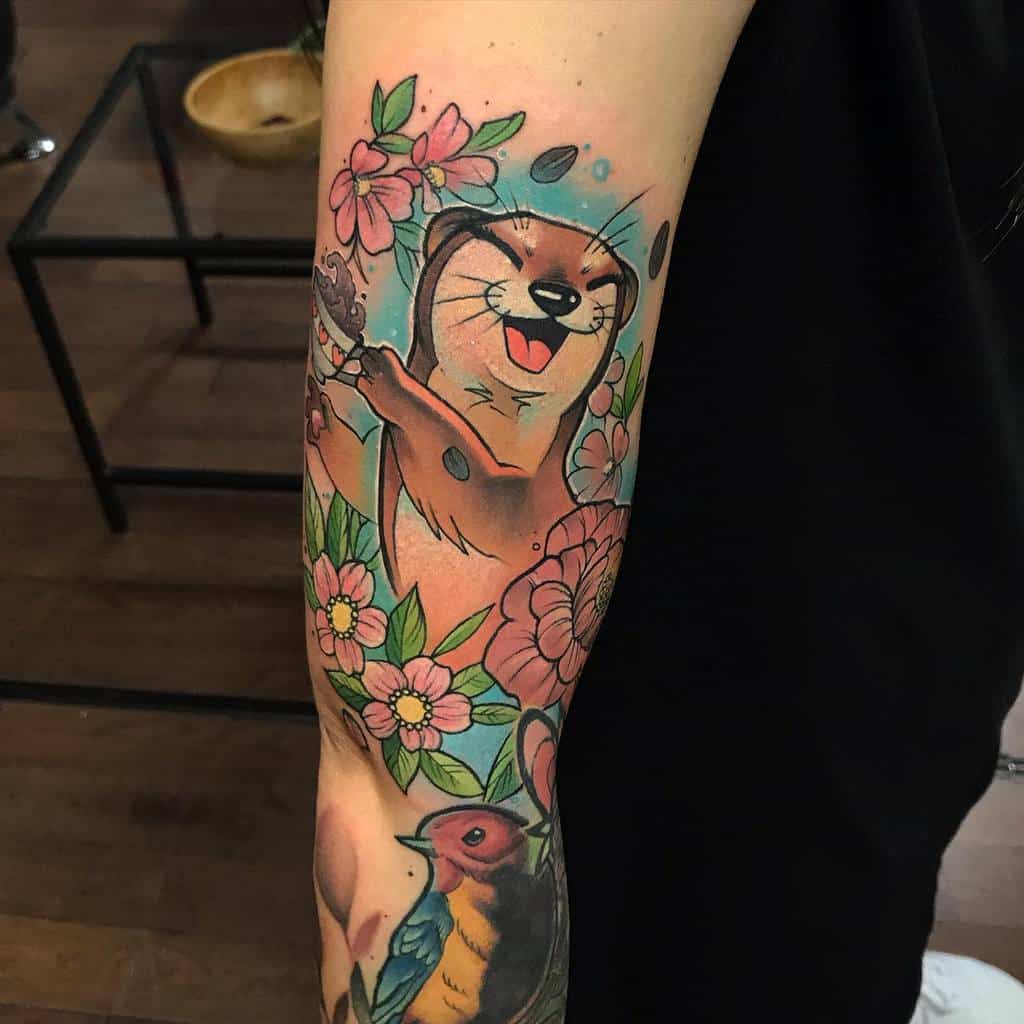 Forearm tattoo of a playful otter with pink flowers and leaves, next to a colorful bird tattoo on an arm