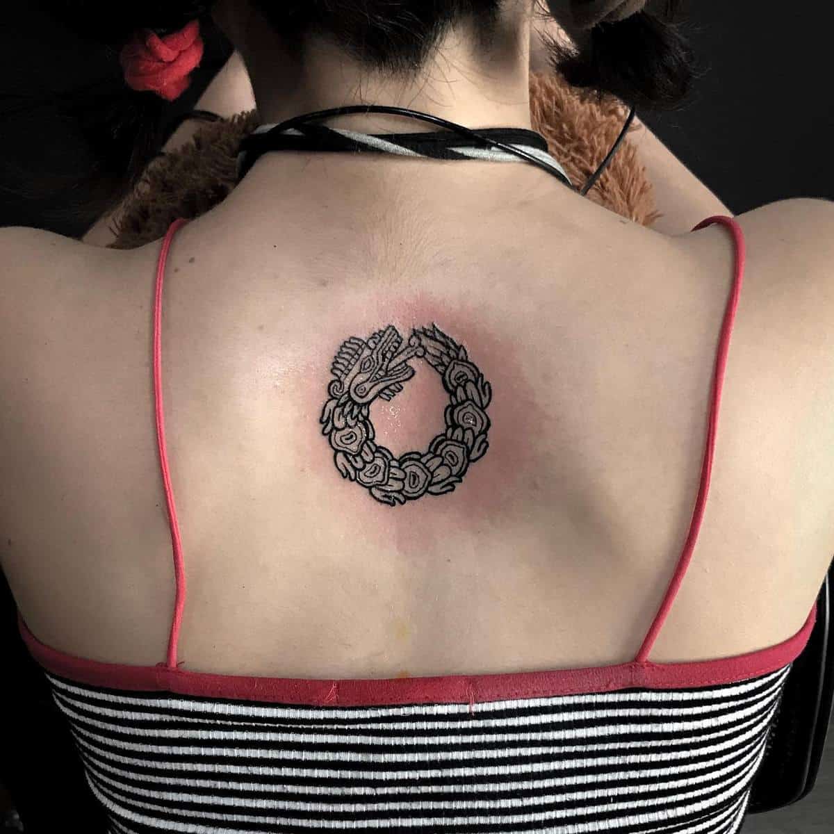 Minimalist black Ouroboros Quetzalcoatl tattoo on the upper back, symbolizing the cycle of life
