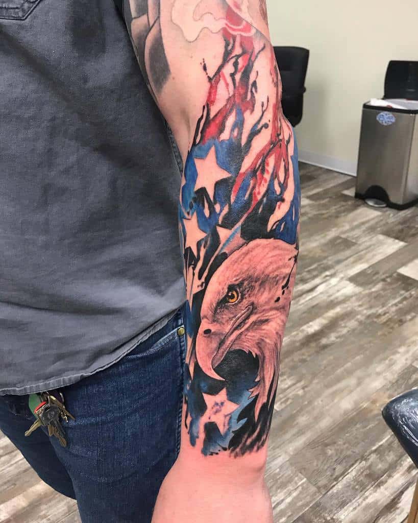 Outer forearm tattoo featuring an American flag design with flowing red, white, and blue colors, stars, and a detailed bald eagle head symbolizing patriotism
