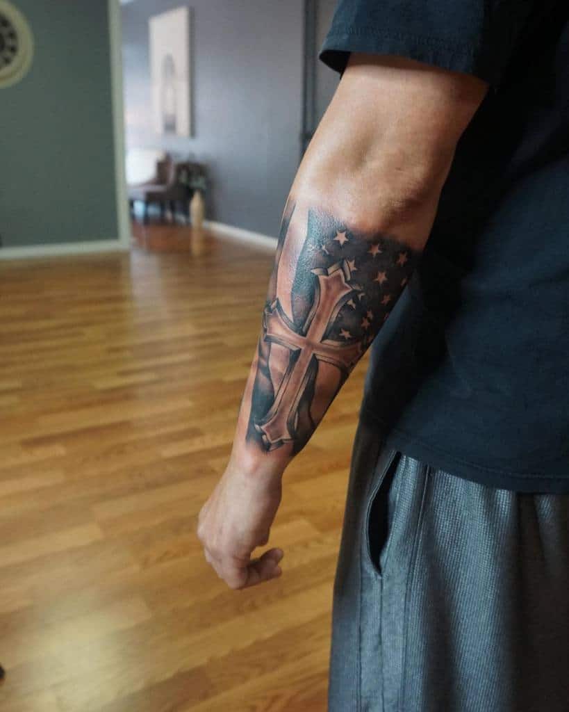 Outer forearm tattoo featuring a cross overlaid on the American flag with stars, symbolizing faith and patriotism