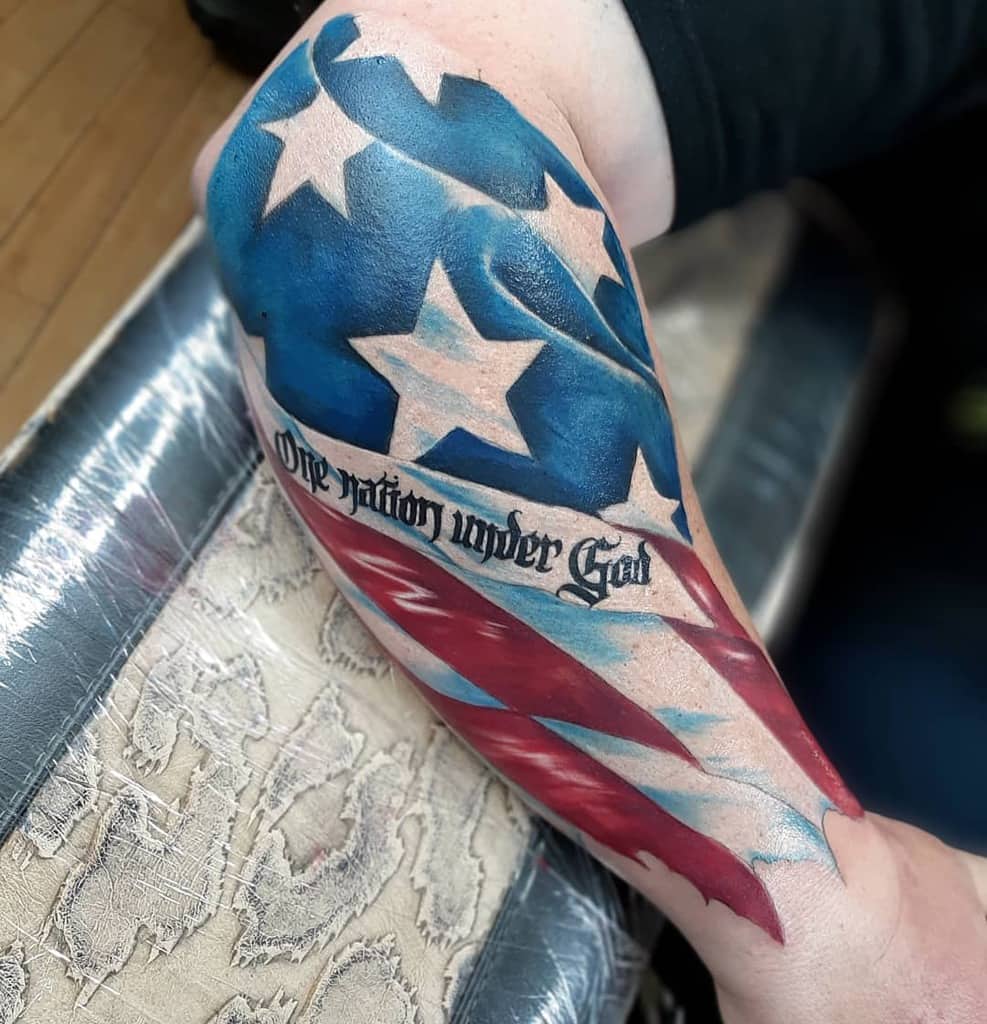 An American flag tattoo with the phrase "One nation under God" elegantly adorns the outer forearm of an arm