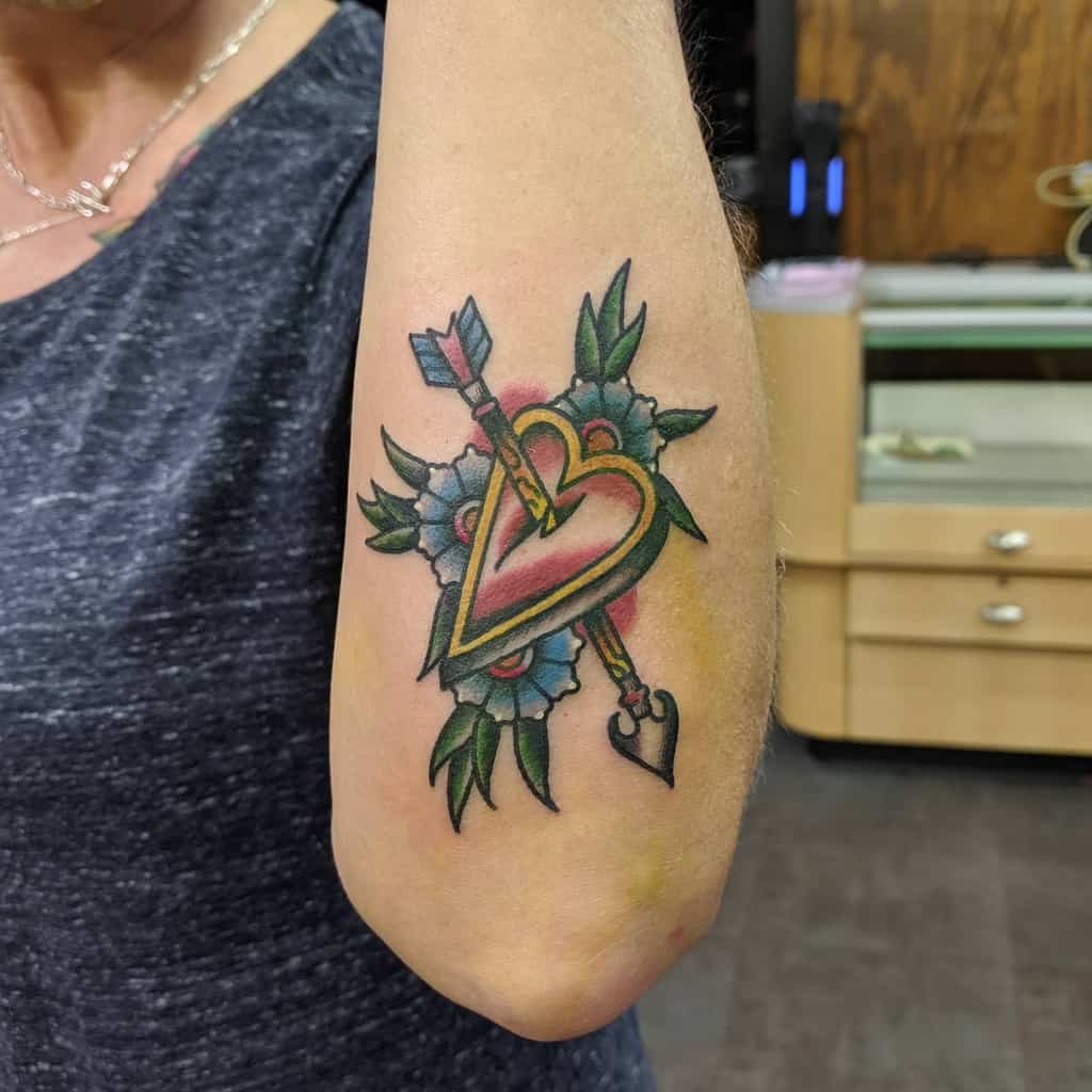 Tattoo of a heart pierced by an arrow, surrounded by flowers, elegantly inked on the outer forearm