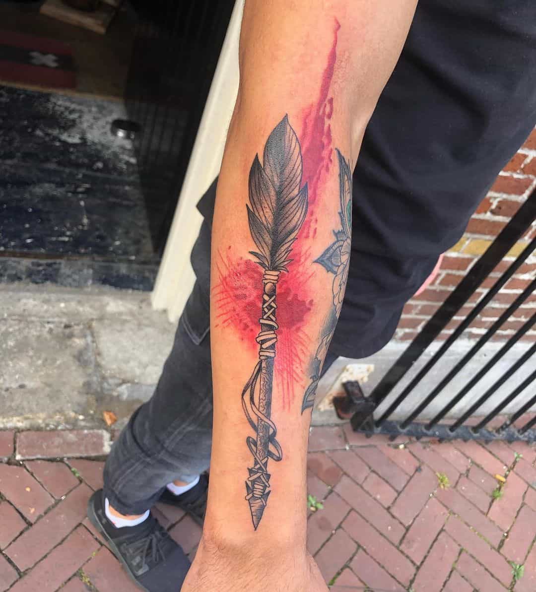 Outer forearm tattoo featuring a detailed arrow with feathers, set against a red watercolor background, symbolizing strength and focus