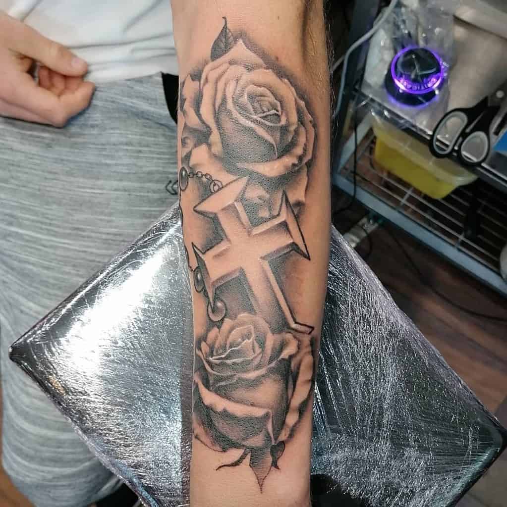 Outer forearm tattoo of a cross intertwined with delicate roses and a graceful rosary