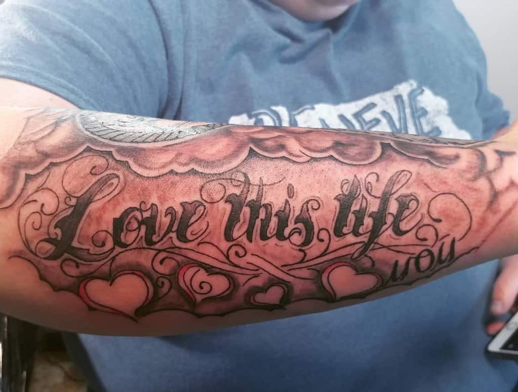Tattooed outer forearm reading "Love this life" adorned with decorative clouds and hearts