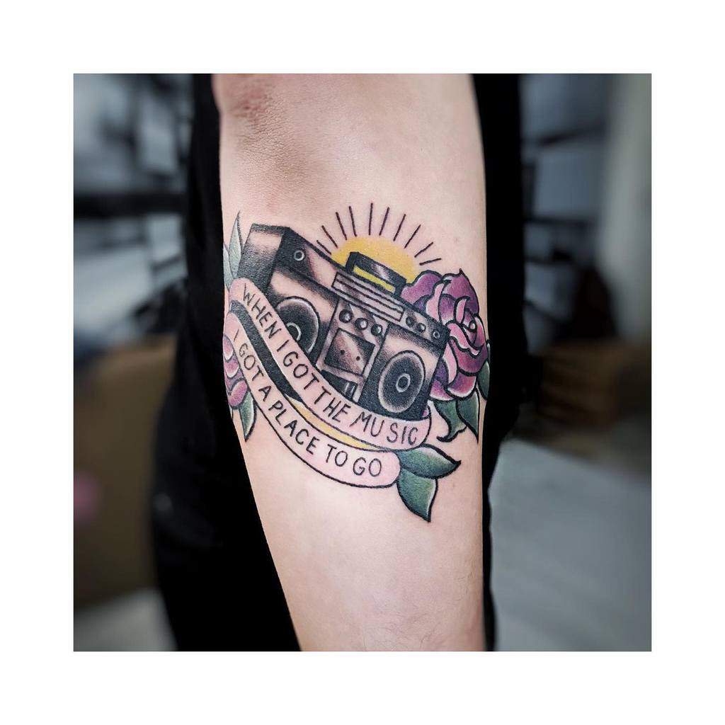 Tattoo on the outer forearm of a camera adorned with flowers and a banner that reads, "When I got the music, I got a place to go"