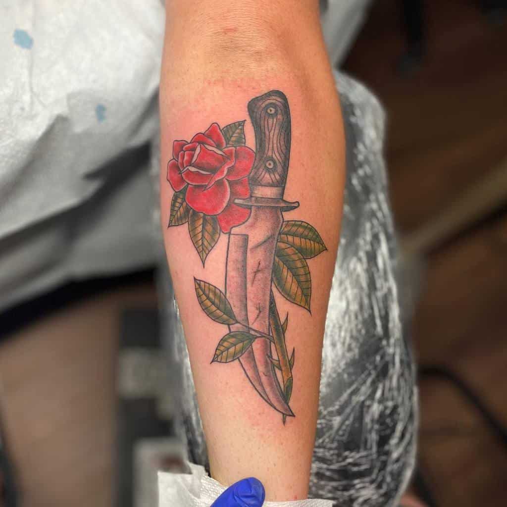 Tattoo of a dagger with a red rose and green leaves on an outer forearm