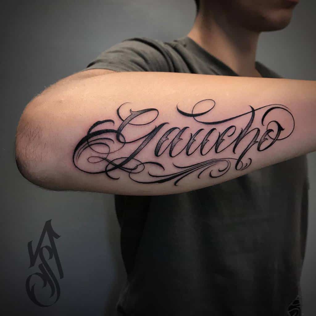 Outer forearm tattoo of the word 'Gaucho' in elegant, flowing script, symbolizing pride in cultural heritage