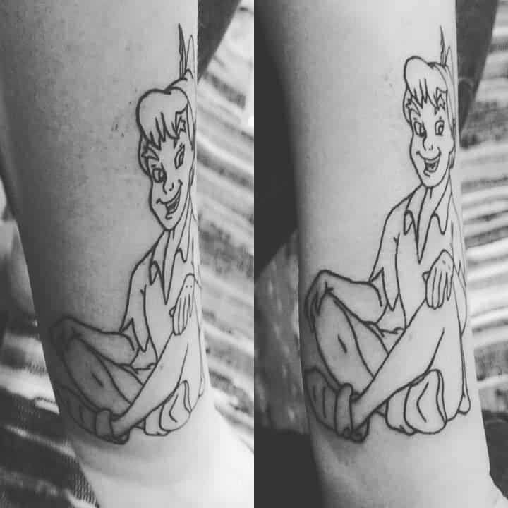 Black line-art tattoo of Peter Pan sitting cross-legged with a playful expression, wearing his signature outfit and feathered hat