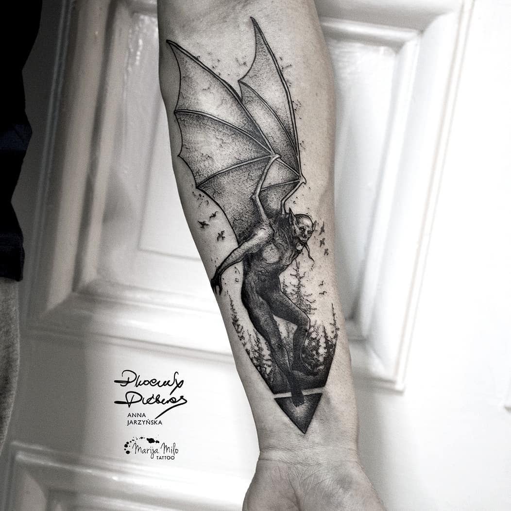 Forearm tattoo with a winged creature, spear-like tail, and surrounding specks