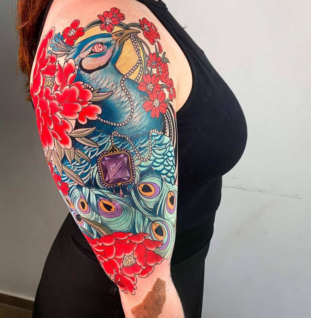 Colorful peacock tattoo with red flowers and a purple gem on upper arm