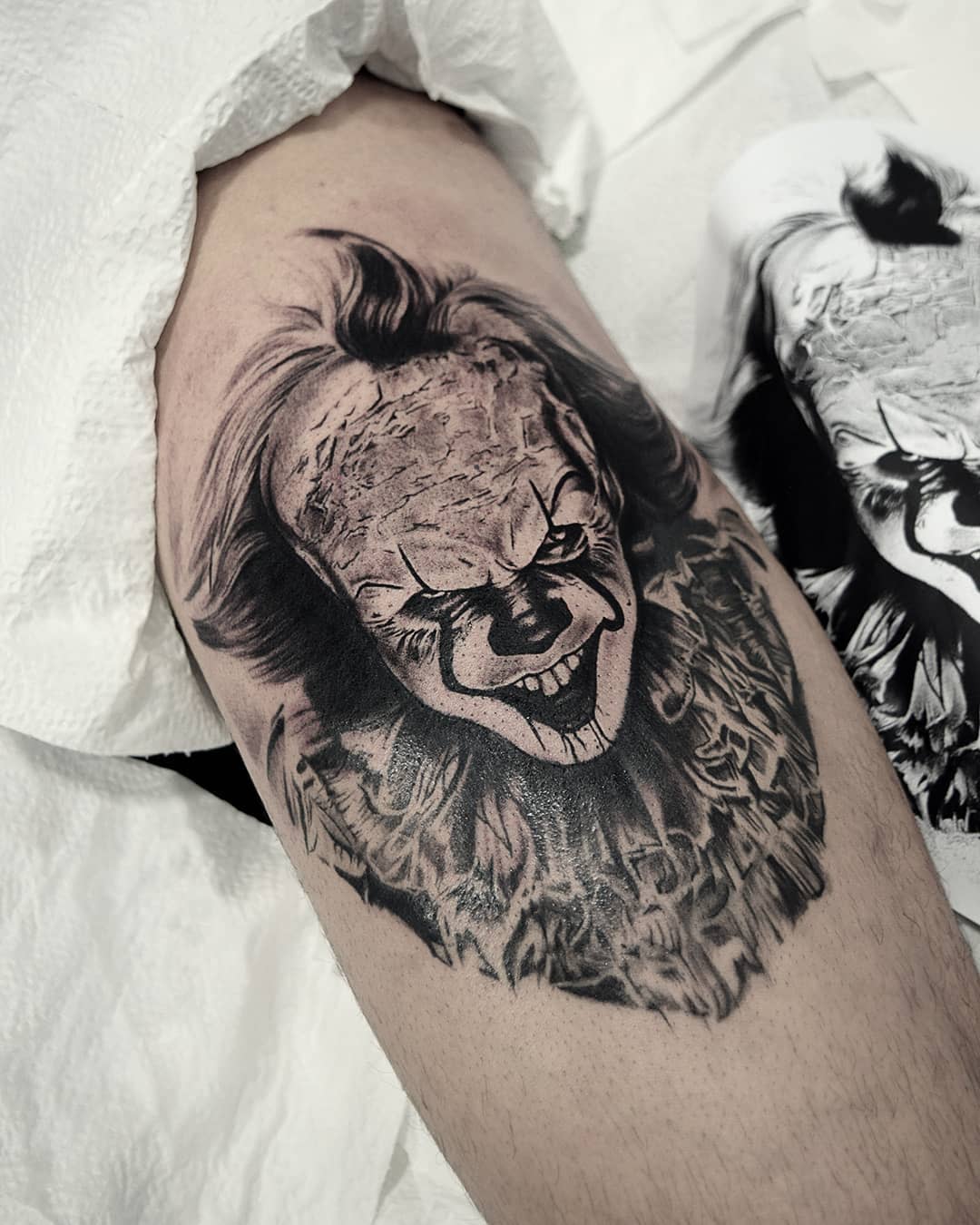 Black and grey Pennywise tattoo on upper arm, showcasing detailed facial expression and hair