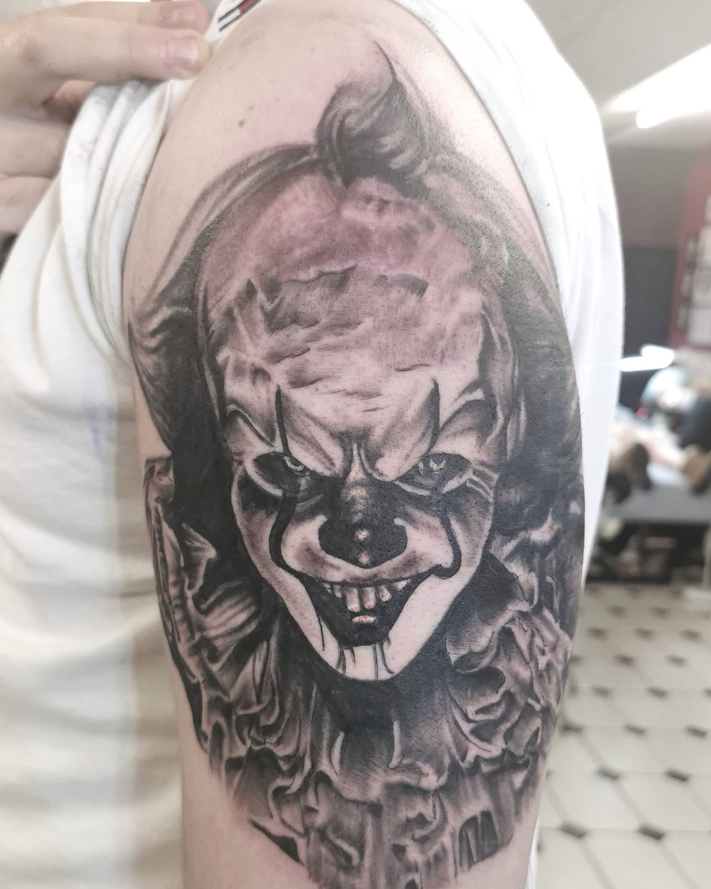 Black and grey Pennywise tattoo on upper arm, emphasizing his sinister smile and intense eyes