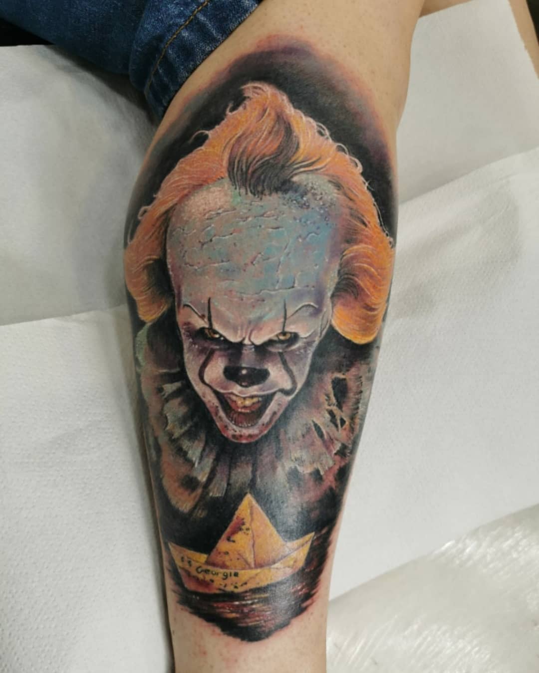 Colorful Pennywise tattoo on calf, featuring his sinister face and Georgie's paper boat