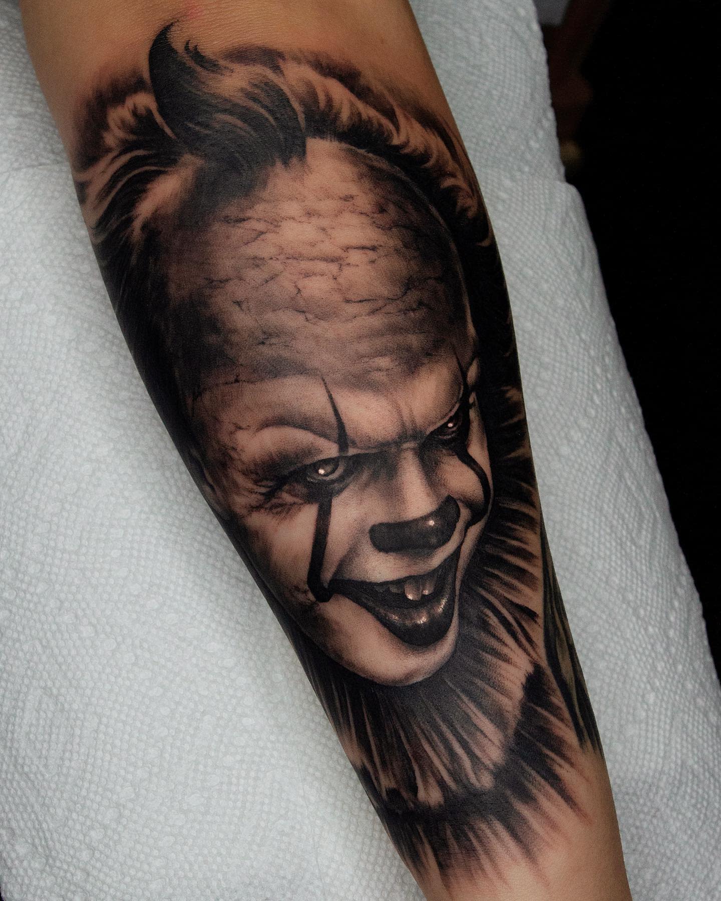 Black and grey Pennywise tattoo on forearm, showcasing his haunting grin and realistic shading