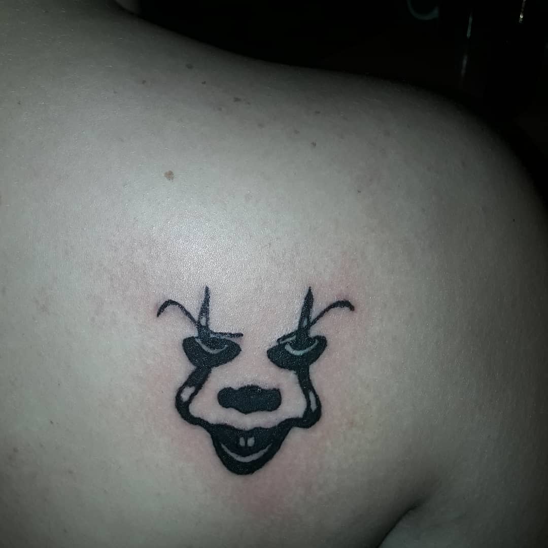 Simple black ink Pennywise face tattoo on shoulder, capturing his eerie expression with minimal lines