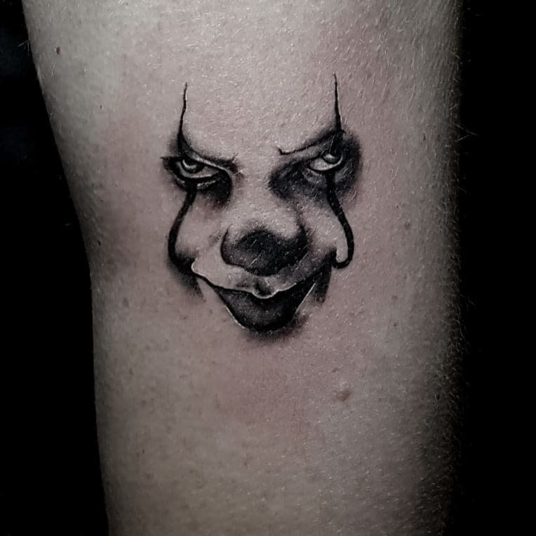 Small black and grey Pennywise face tattoo on leg, featuring intense shading and a sinister expression