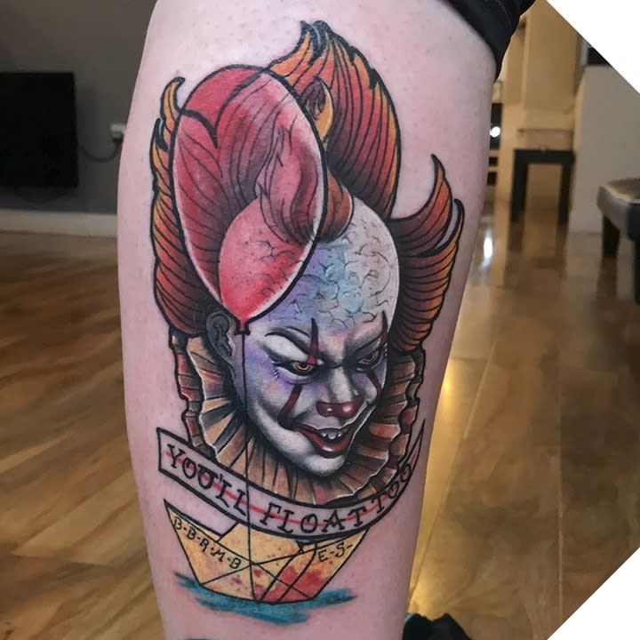A traditional-style Pennywise tattoo featuring his face, a red balloon, and the phrase "You'll Float Too" above a paper boat