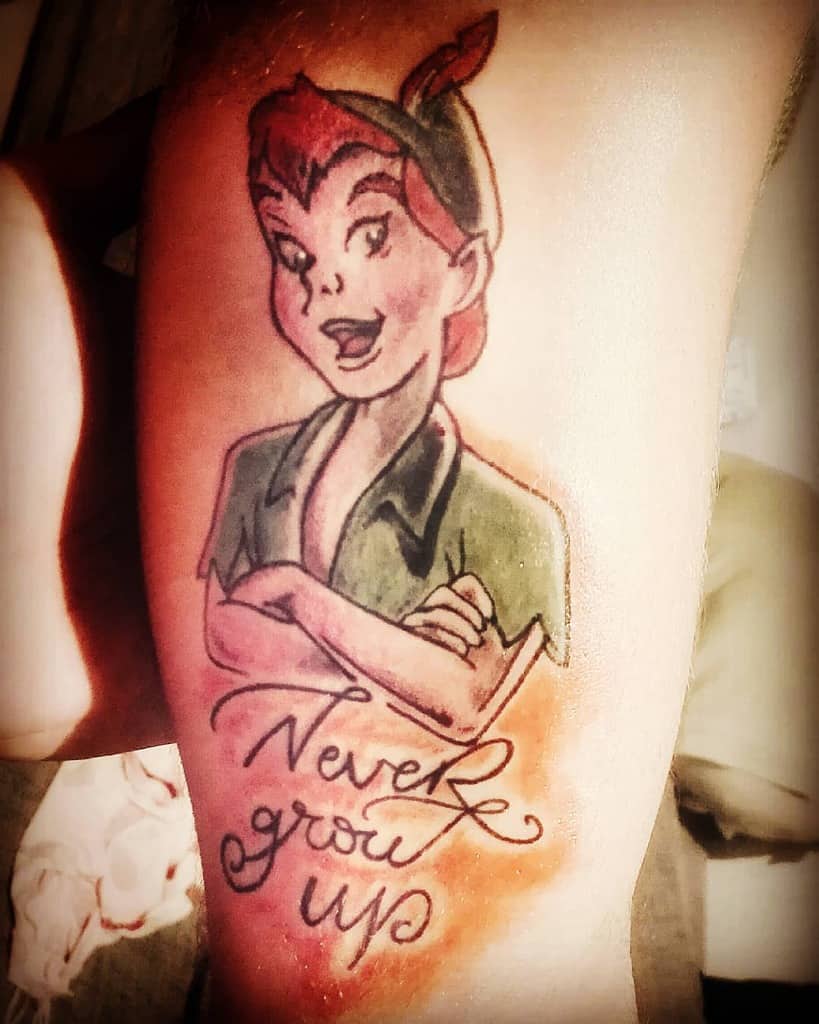 Tattoo of Peter Pan with a playful expression, arms crossed, wearing his signature green outfit and hat, with the phrase "Never Grow Up"