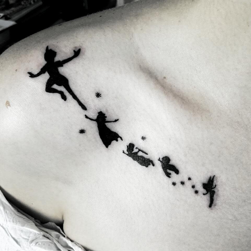 Peter Pan, Wendy, John, and Michael flying with Tinker Bell, leaving a trail of stars in a minimalist black ink tattoo