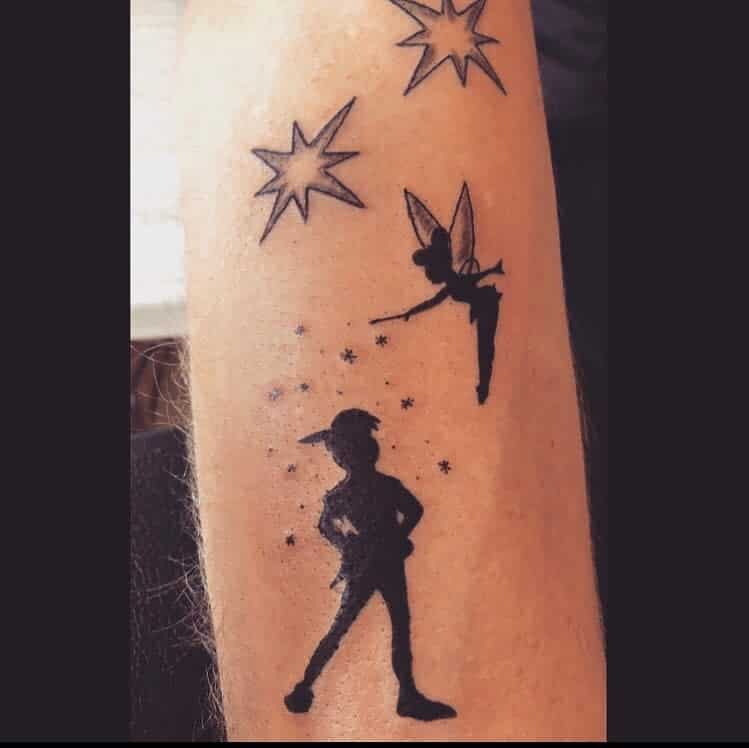 Black silhouette tattoo of Peter Pan standing confidently, with Tinker Bell sprinkling pixie dust above, surrounded by stars