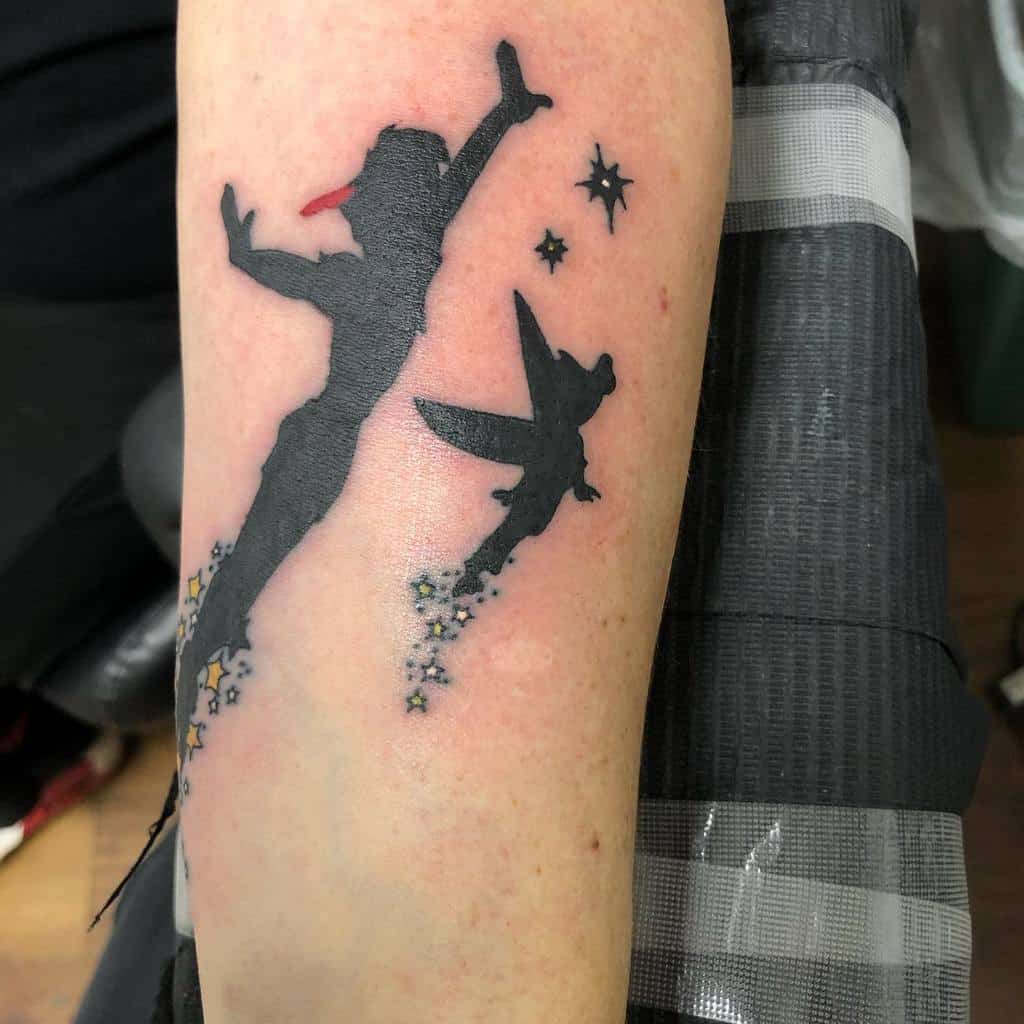 Black silhouette tattoo of Peter Pan and Tinker Bell flying among stars, with sparkling pixie dust trailing