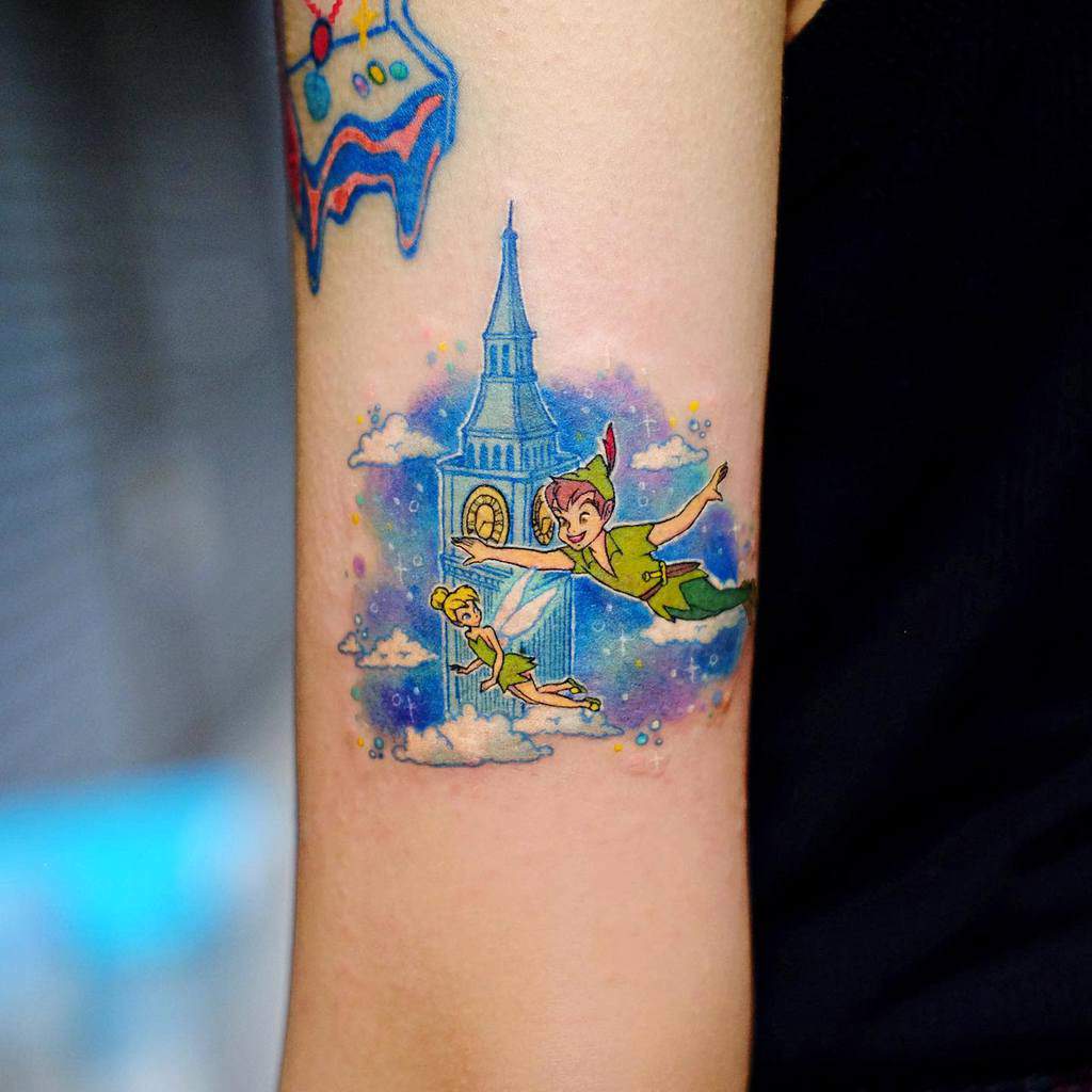 Colorful tattoo of Peter Pan and Tinker Bell flying past Big Ben, surrounded by fluffy clouds and a dreamy blue watercolor background