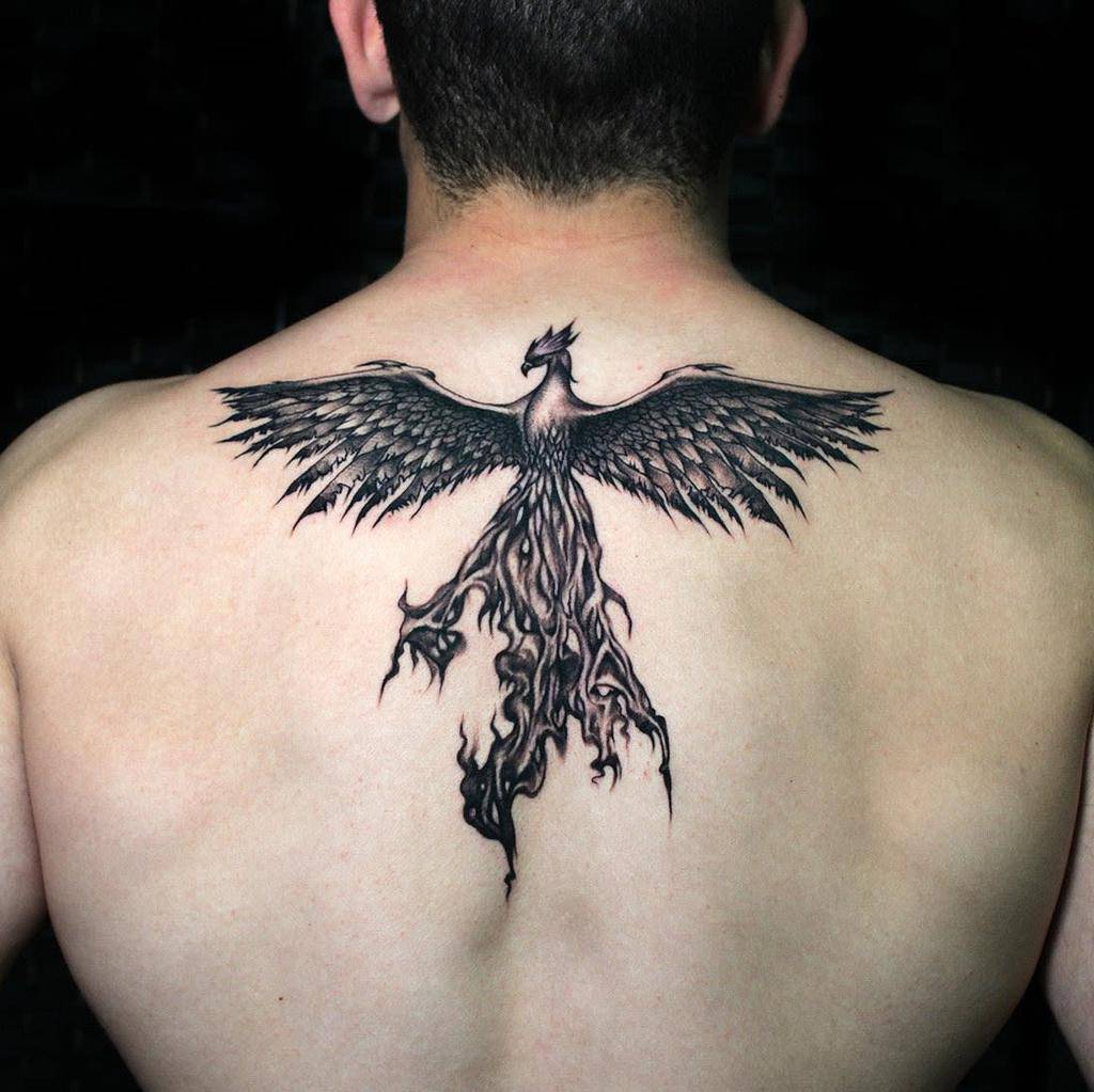 A person has a detailed phoenix rising tattoo on their upper back with wings spread wide